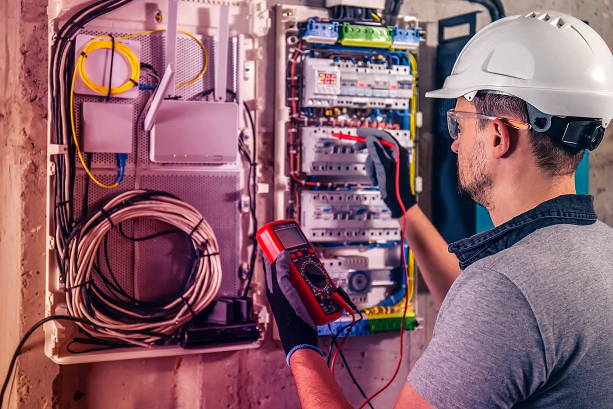 Baretli elektrik ustası, açık dağıtım panosunda ölçü aletiyle sigortaları ölçüyor